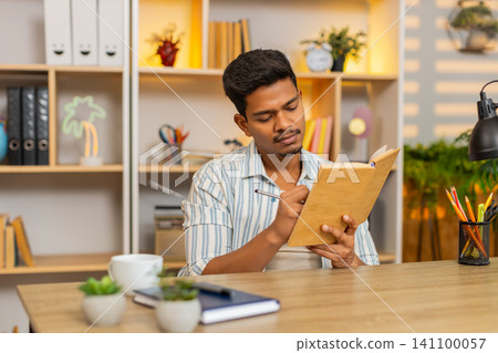 Indian man on home table reading book making notes and thinking deeply during focused study at home Indian man on home table reading book making notes and thinking deeply during focused study at home 141100057
