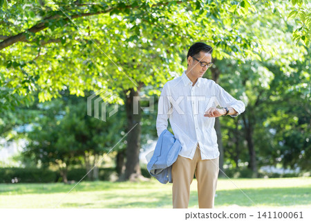 A senior man wearing glasses takes a walk in a park surrounded by fresh greenery. A senior man wearing glasses takes a walk in a park surrounded by fresh greenery. 141100061
