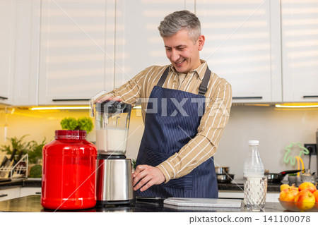 Middle-aged man making protein shake in kitchen with blender for fitness nutrition and home routine Middle-aged man making protein shake in kitchen with blender for fitness nutrition and home routine 141100078