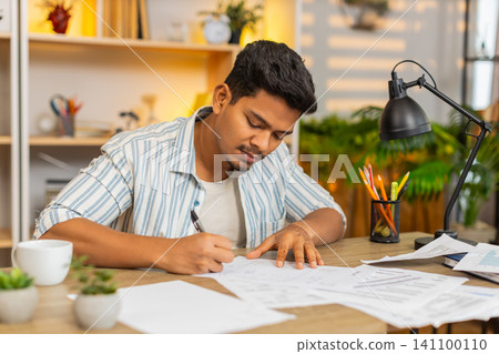 Indian man on home table reading final line on paper before signing office document before approval Indian man on home table reading final line on paper before signing office document before approval 141100110