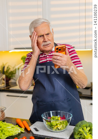 Senior old man upset reading bad news on smartphone in kitchen at home facing bank scam alert stress Senior old man upset reading bad news on smartphone in kitchen at home facing bank scam alert stress 141100239