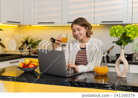 Young woman drinking orange juice while using laptop in kitchen for healthy eating and online chat Young woman drinking orange juice while using laptop in kitchen for healthy eating and online chat 141100379