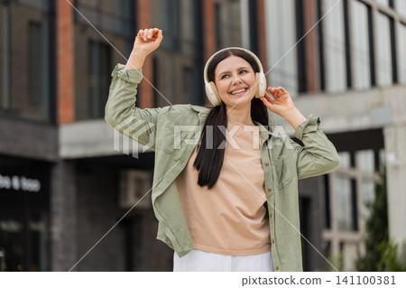 Young woman listening to music walking in city street smiling dancing with headphones during stroll 141100381