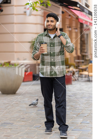 Indian man listening to music walking in city street with wireless headphones smiling during stroll Indian man listening to music walking in city street with wireless headphones smiling during stroll 141100838