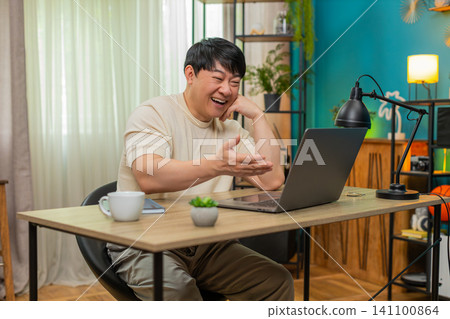 Asian man smiling watching nostalgic video on laptop, resting head on hand, enjoying warm memories Asian man smiling watching nostalgic video on laptop, resting head on hand, enjoying warm memories 141100864