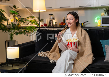 Young woman wrapped in plaid holds popcorn watches comedy on tv laughs comments comfy cozy evening 141100873