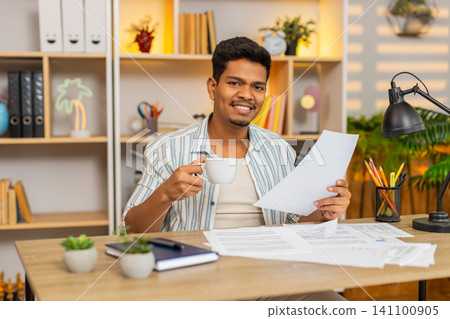 Indian man on home table reviewing work documents and sipping coffee during focused office routine Indian man on home table reviewing work documents and sipping coffee during focused office routine 141100905