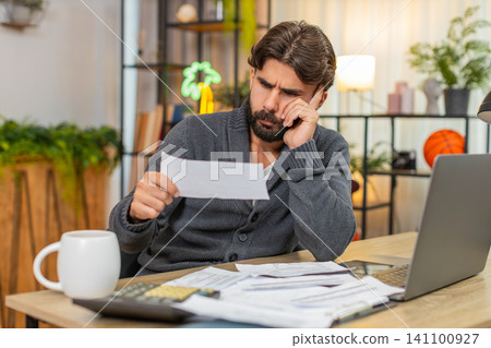 Indian man bends forward head in hands unpaid bills on table calculator and laptop heavy worry money Indian man bends forward head in hands unpaid bills on table calculator and laptop heavy worry money 141100927