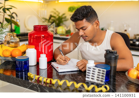 Indian man making sports supplement shopping list for fitness nutrition routine in kitchen at home Indian man making sports supplement shopping list for fitness nutrition routine in kitchen at home 141100931
