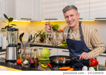 Middle-aged man smiling while stirring vegetables in frying pan in kitchen at home for healthy meal Middle-aged man smiling while stirring vegetables in frying pan in kitchen at home for healthy meal 141100959