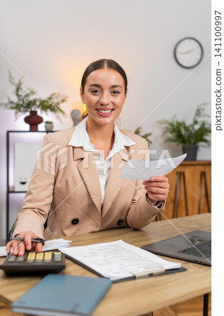 Young businesswoman totals month on calculator checks receipts and invoices nods with satisfaction 141100997