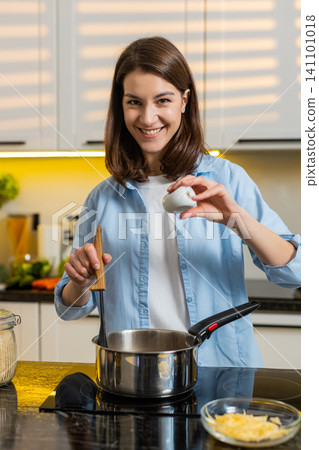 Young woman seasoning soup on stove in home kitchen smiling while cooking healthy diet recipe dinner Young woman seasoning soup on stove in home kitchen smiling while cooking healthy diet recipe dinner 141101018