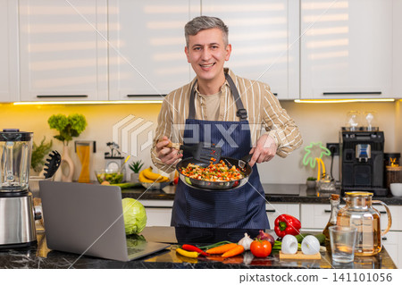Middle-aged man frying vegetables while reading laptop screen for healthy diet meal in home kitchen Middle-aged man frying vegetables while reading laptop screen for healthy diet meal in home kitchen 141101056