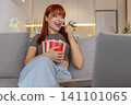 Young woman relaxes with popcorn watches funny trailer on laptop laughs smiles enjoys comedy preview 141101065