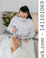 Adult woman receives rejuvenating facial therapy in cosmetology clinic by professional cosmetologist 141101069