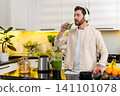 Middle-aged man drinking green smoothie wearing headphones in kitchen for healthy diet and fitness 141101078