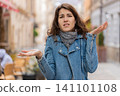 Young woman showing confusion while walking in city street with puzzled look and questioning gesture 141101108