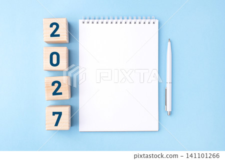 Wooden cubes with numbers for the New Year 2027 with a notebook and pen on a blue background Wooden cubes with numbers for the New Year 2027 with a notebook and pen on a blue background 141101266