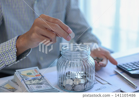 Saving Money Concept. Businessman Putting Coins into Glass Jar Smart Money Habits and Financial Literacy Visual Saving Money Concept. Businessman Putting Coins into Glass Jar Smart Money Habits and Financial Literacy Visual 141105486