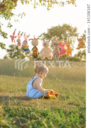 Soft children's toys are hanging to dry on a clothesline, and a baby is sitting on the grass, playing with a stuffed bull. Soft children's toys are hanging to dry on a clothesline, and a baby is sitting on the grass, playing with a stuffed bull. 141106167