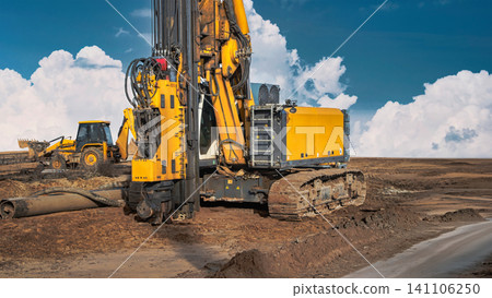 A drilling rig operates on a construction site, driving piles into the ground. Nearby, a bulldozer prepares the area. Clear skies provide good visibility A drilling rig operates on a construction site, driving piles into the ground. Nearby, a bulldozer prepares the area. Clear skies provide good visibility 141106250