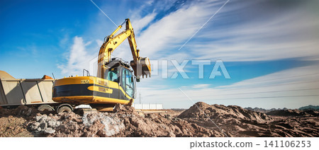 An excavator lifts dirt at a construction site. The work is part of a larger project in a dry area. Trucks are nearby to transport materials 141106253