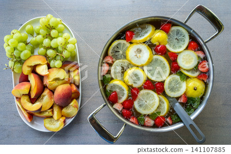 Fresh fruit assortment with grapes and sliced peaches beside citrus strawberry infused water in stainless pot on gray tabletop, bright healthy summer food presentation from overhead Fresh fruit assortment with grapes and sliced peaches beside citrus strawberry infused water in stainless pot on gray tabletop, bright healthy summer food presentation from overhead 141107885