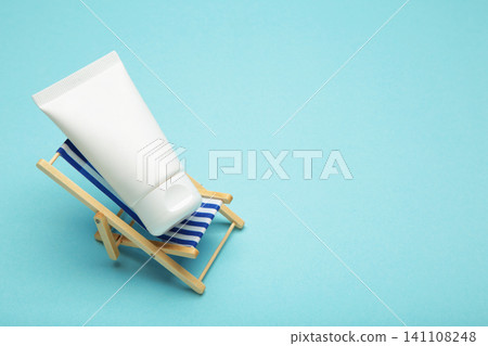 Cosmetic facial cream in plastic tube on miniature deck chair on blue background. Summer concept. Cosmetic facial cream in plastic tube on miniature deck chair on blue background. Summer concept. 141108248