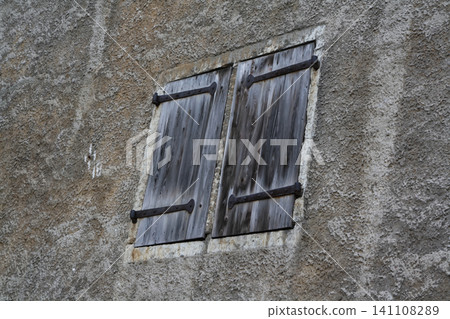 Close-Up Architectural View of Weathered Grey Wooden Shutters with Wrought Iron Hinges on a Rough Textured Plaster Wall 141108289
