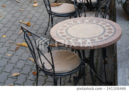 Charming Outdoor Cafe Seating Featuring a Mosaic Tiled Round Table and Decorative Wrought Iron Chairs on an Autumn Sidewalk 141108466