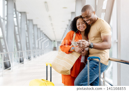 Happy young Black couple standing close in sunny airport corridor, checking smartphone Happy young Black couple standing close in sunny airport corridor, checking smartphone 141108855