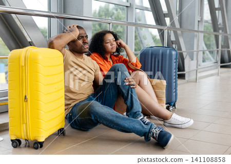 Young Black couple sitting on airport corridor floor beside suitcases, looking tired Young Black couple sitting on airport corridor floor beside suitcases, looking tired 141108858