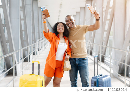 Black couple raising passports and boarding passes in airport corridor with suitcases 141108914