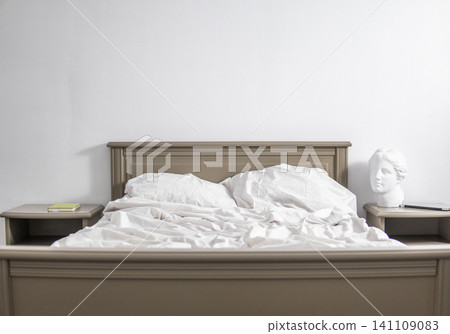 Unmade bed in a minimalist bedroom with white wall and classical bust, symbolizing rest and morning routine Unmade bed in a minimalist bedroom with white wall and classical bust, symbolizing rest and morning routine 141109083