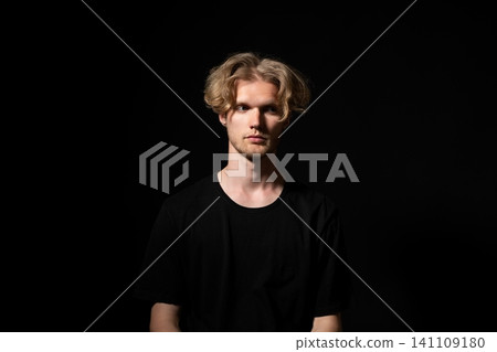 Young man with blond hair and serious expression posing in studio with dramatic lighting and black background, symbolizing contemplation, depth, and introspection 141109180