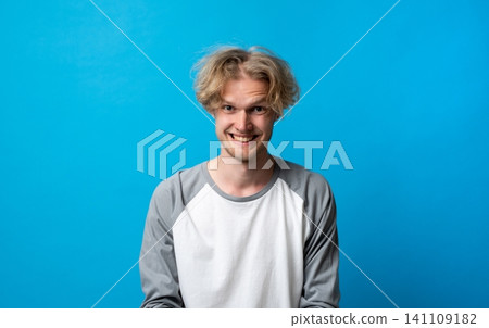Young man with messy blond hair and a mischievous grin, posing for a cropped studio headshot against a blue backdrop, playful, confident and candid expression Young man with messy blond hair and a mischievous grin, posing for a cropped studio headshot against a blue backdrop, playful, confident and candid expression 141109182