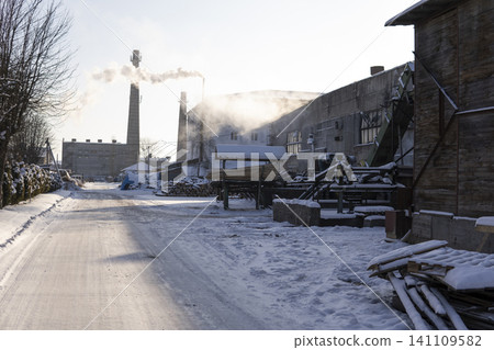 Snow-covered industrial factory operating in winter, showing smoking chimneys, a raw wood processing area, and machinery Snow-covered industrial factory operating in winter, showing smoking chimneys, a raw wood processing area, and machinery 141109582