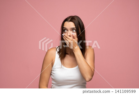 Young woman covering mouth in shock and wide-eyed surprise, expressing emotion on pink background 141109599