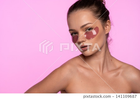 Young woman wearing pink eye patches for under eye skincare during beauty routine on pink background Young woman wearing pink eye patches for under eye skincare during beauty routine on pink background 141109687