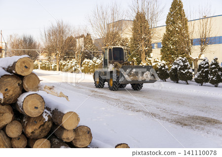 Front loader clearing snow from a road in front of a factory building, with wooden logs covered in snow stacked nearby Front loader clearing snow from a road in front of a factory building, with wooden logs covered in snow stacked nearby 141110078
