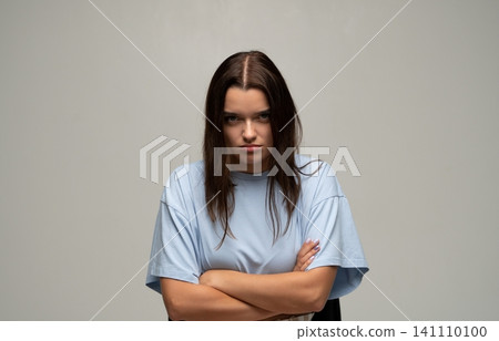 Frowning young woman standing with folded arms, her expression conveying a mix of anger, annoyance, and bitterness 141110100