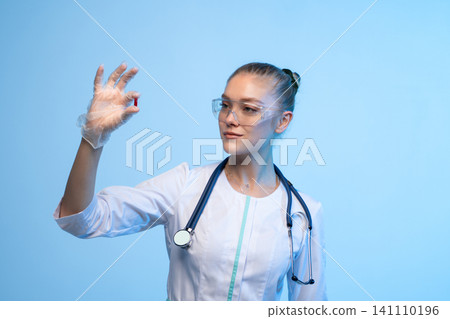 Female doctor in lab coat and safety goggles observing a red capsule, representing medical research or treatment Female doctor in lab coat and safety goggles observing a red capsule, representing medical research or treatment 141110196