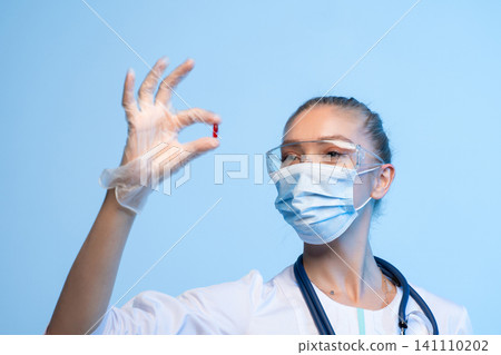 Medical professional wearing mask and safety glasses holding a red capsule, representing health, medicine, and research developing treatment Medical professional wearing mask and safety glasses holding a red capsule, representing health, medicine, and research developing treatment 141110202