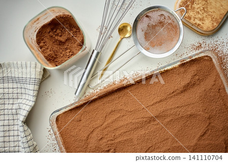 Cocoa covered tiramisu sits in rectangular glass dish beside sieve and cocoa bowl on table Cocoa covered tiramisu sits in rectangular glass dish beside sieve and cocoa bowl on table 141110704