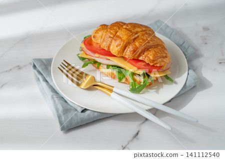 A golden croissant sandwich with ham, cheese, tomato, and arugula on a marble background A golden croissant sandwich with ham, cheese, tomato, and arugula on a marble background 141112540