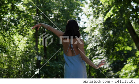 Graceful woman seen from behind in light blue dress dancing with raised arms among lush green trees, soft sunlight filtering through foliage, calm summer park mood and natural 141113103
