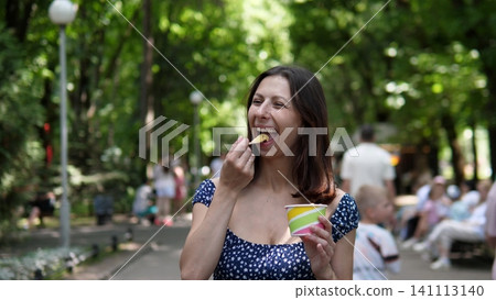 A woman is happily enjoying ice cream in a park, surrounded by trees and people. She is wearing a cloth dress on a sunny summer day with a blue sky and white clouds. A woman is happily enjoying ice cream in a park, surrounded by trees and people. She is wearing a cloth dress on a sunny summer day with a blue sky and white clouds. 141113140