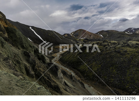 Dramatic volcanic canyon with a winding glacial river, colorful rhyolite mountains, lava fields and snow patches beneath storm clouds in Landmannalaugar, Iceland Highlands. Dramatic volcanic canyon with a winding glacial river, colorful rhyolite mountains, lava fields and snow patches beneath storm clouds in Landmannalaugar, Iceland Highlands. 141113296