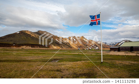 Icelandic flag waving in the wind against the colorful rhyolite mountains of Landmannalaugar in the Iceland Highlands under dramatic clouds and evening light. 141113298