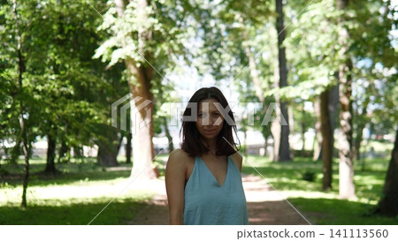 Serene outdoor portrait showing young woman with dark hair in sleeveless blue dress standing on shaded path among tall trees, soft daylight, blurred leafy background 141113560